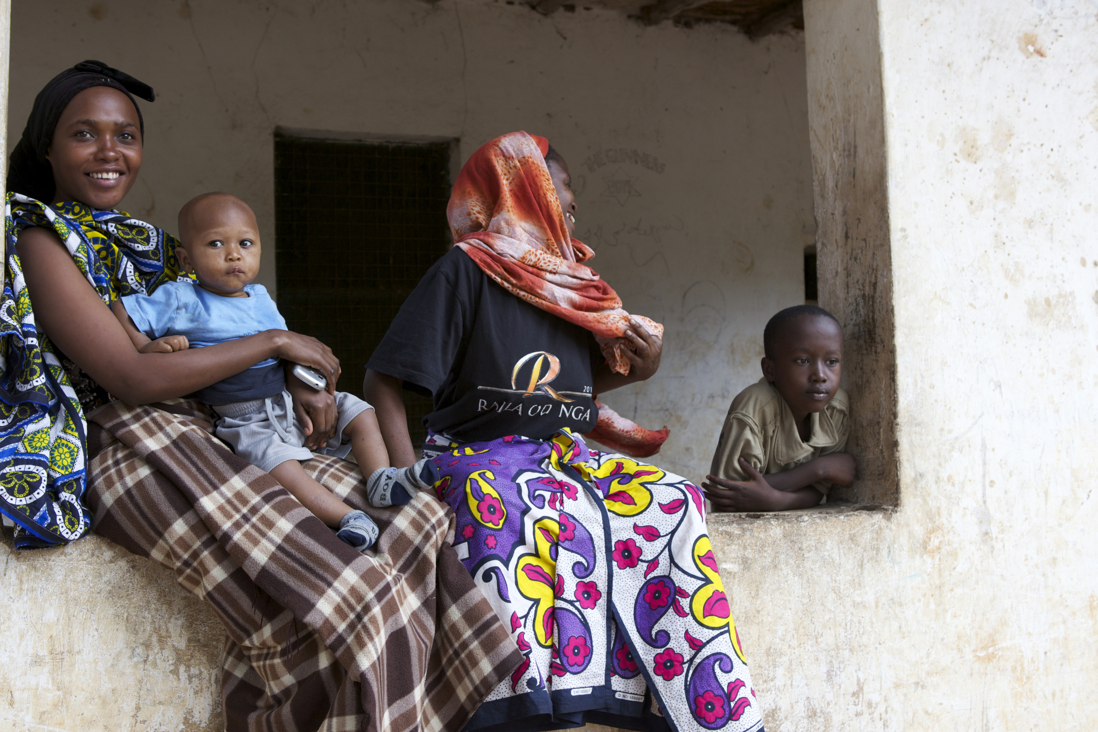 Two women sitting on the ledge of a window cut into a stucco wall, one woman is holding a baby and the other looks back towards a young boy leaning on the ledge.